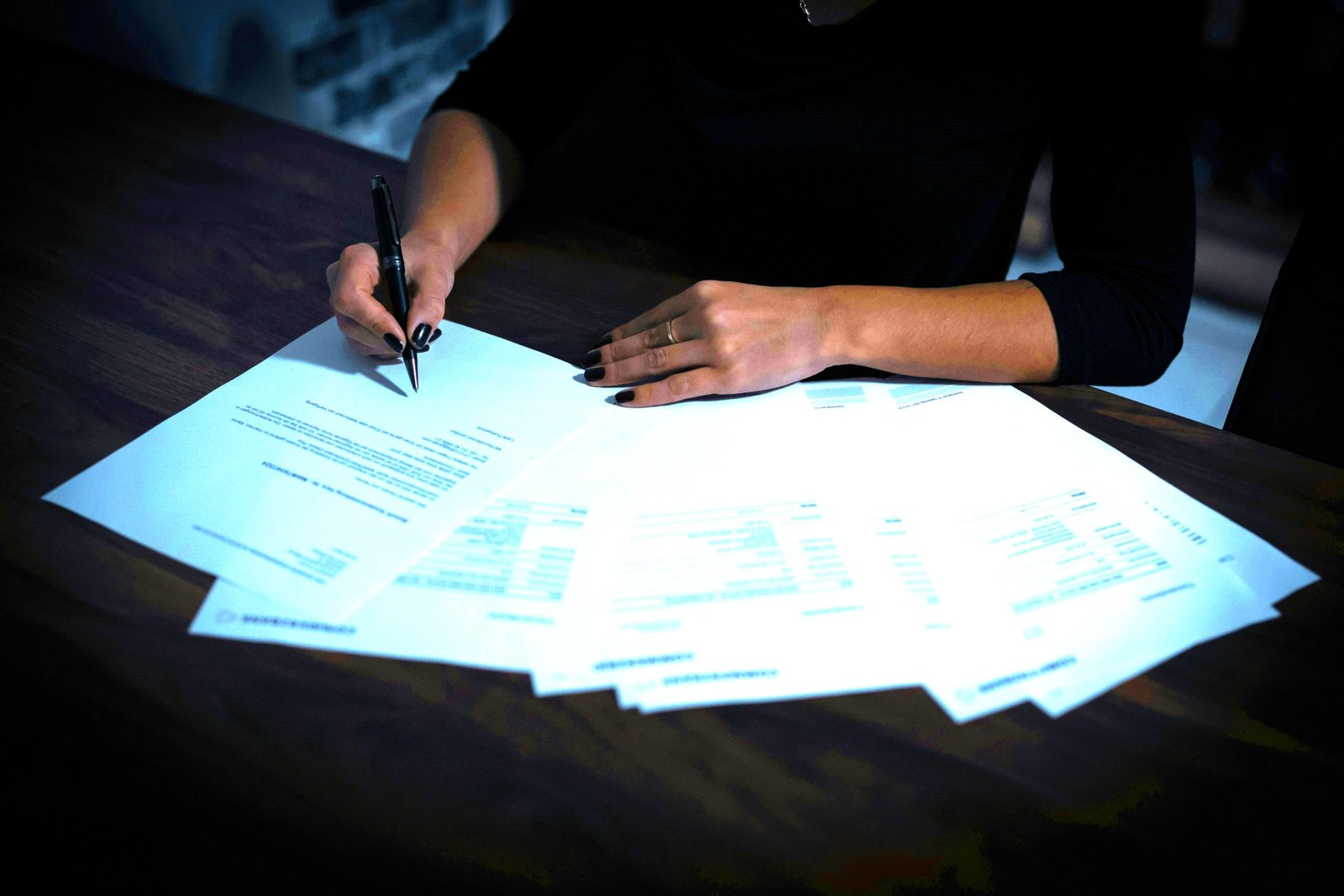 Une femme tient un stylo au dessus d'une liasse de documents disposés sur une table marron.